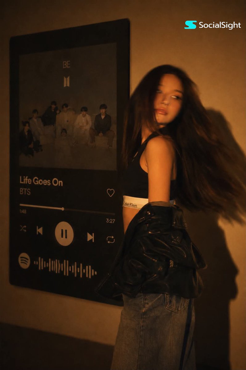一位发型飘逸的深色长发时尚年轻女子在巨型壁挂Spotify音乐播放器显示屏前摆姿势。温暖的琥珀色电影级灯光布满房间,在墙上投下戏剧性阴影。她身穿黑色卡尔文·克莱因露脐上衣,廓形黑…