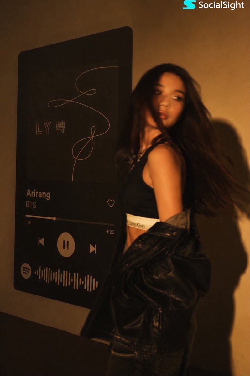 一位发型飘逸的深色长发时尚年轻女子在巨型壁挂Spotify音乐播放器显示屏前摆姿势。温暖的琥珀色电影级灯光布满房间,在墙上投下戏剧性阴影。她身穿黑色卡尔文·克莱因露脐上衣,廓形黑…