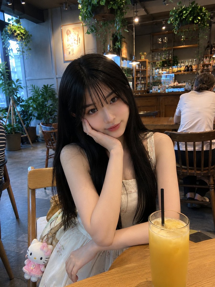 年轻的日本女孩，留着柔顺的长黑发、轻柔蓬松的刘海、白皙透亮的肌肤和一双大而传神的眼睛，随意地坐在一家温馨现代的咖啡馆里。身穿一件精致的奶油白无袖连衣裙，裙身点缀着细腻的花卉图案…