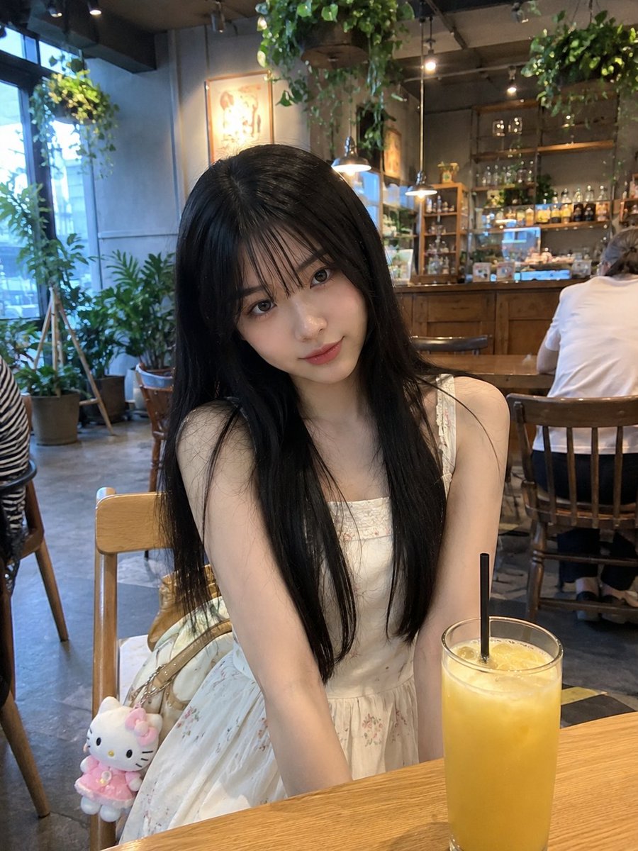 年轻的日本女孩，留着柔顺的长黑发、轻柔蓬松的刘海、白皙透亮的肌肤和一双大而传神的眼睛，随意地坐在一家温馨现代的咖啡馆里。身穿一件精致的奶油白无袖连衣裙，裙身点缀着细腻的花卉图案…