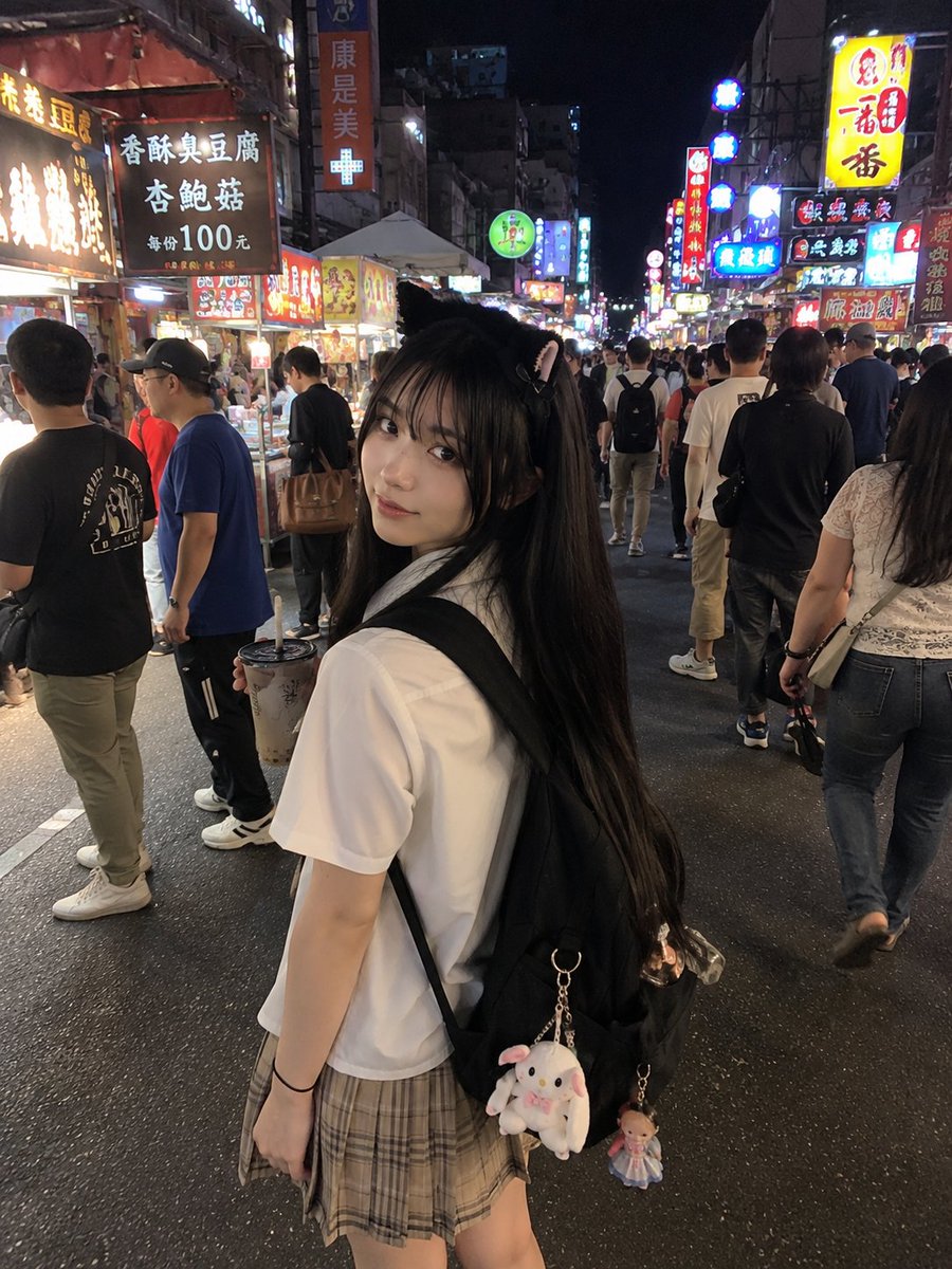 年轻的日本女孩，留着顺直黑色长发和柔软的刘海，戴着可爱的黑色猫耳发箍，身穿白色短袖衬衫、条纹领带和米色格纹百褶裙。她站在一个充满活力的亚洲夜市里，四周是霓虹招牌、美食摊位和熙熙攘…