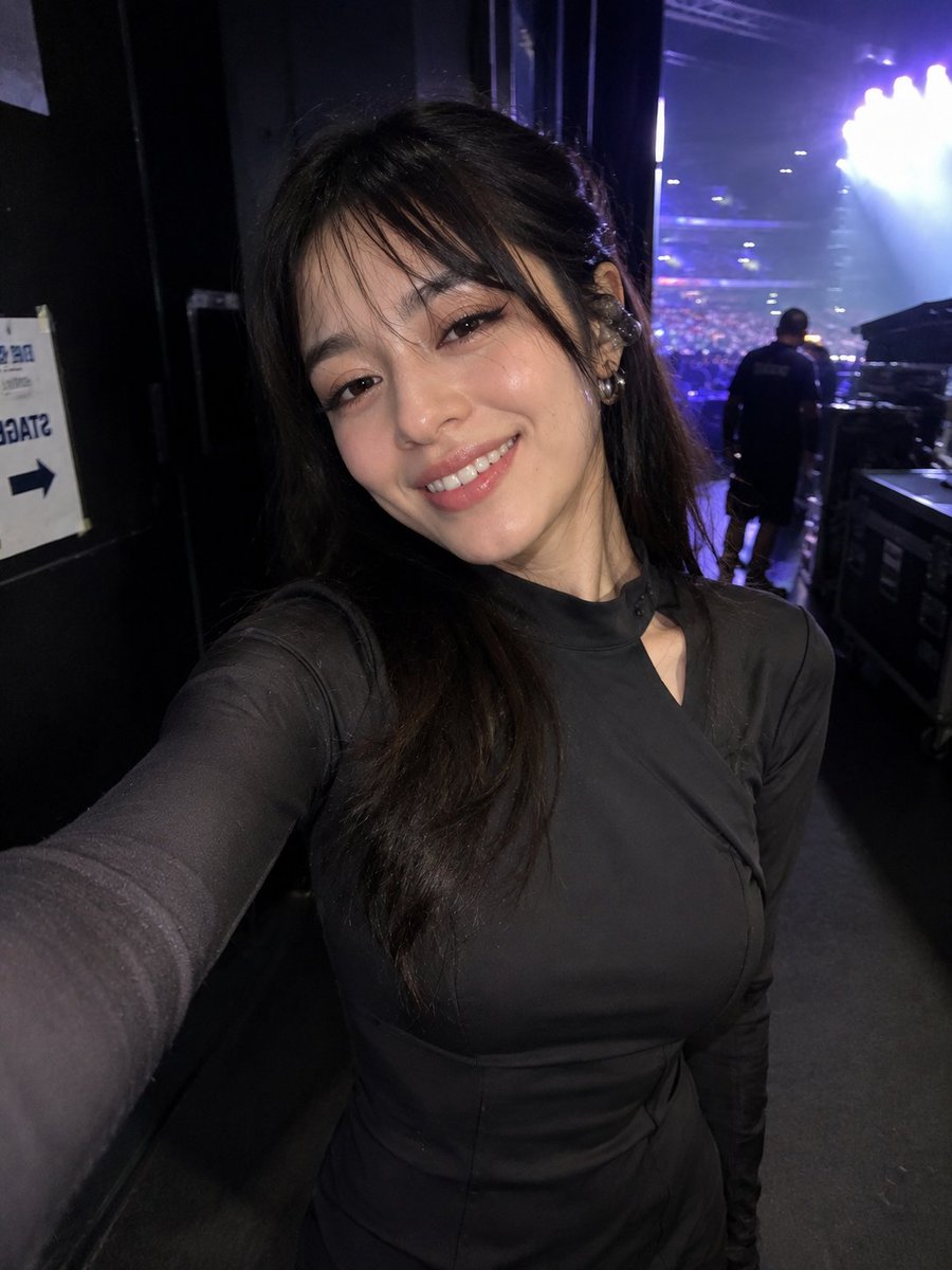 参考图片中同一女性的超写实偶然iPhone前置摄像头自拍,保留完全一致的身份、面部特征、发型和比例。在一场现场演唱会的后台,在她走过走廊时以抓拍方式捕捉。她微微转身,带着放松、演…