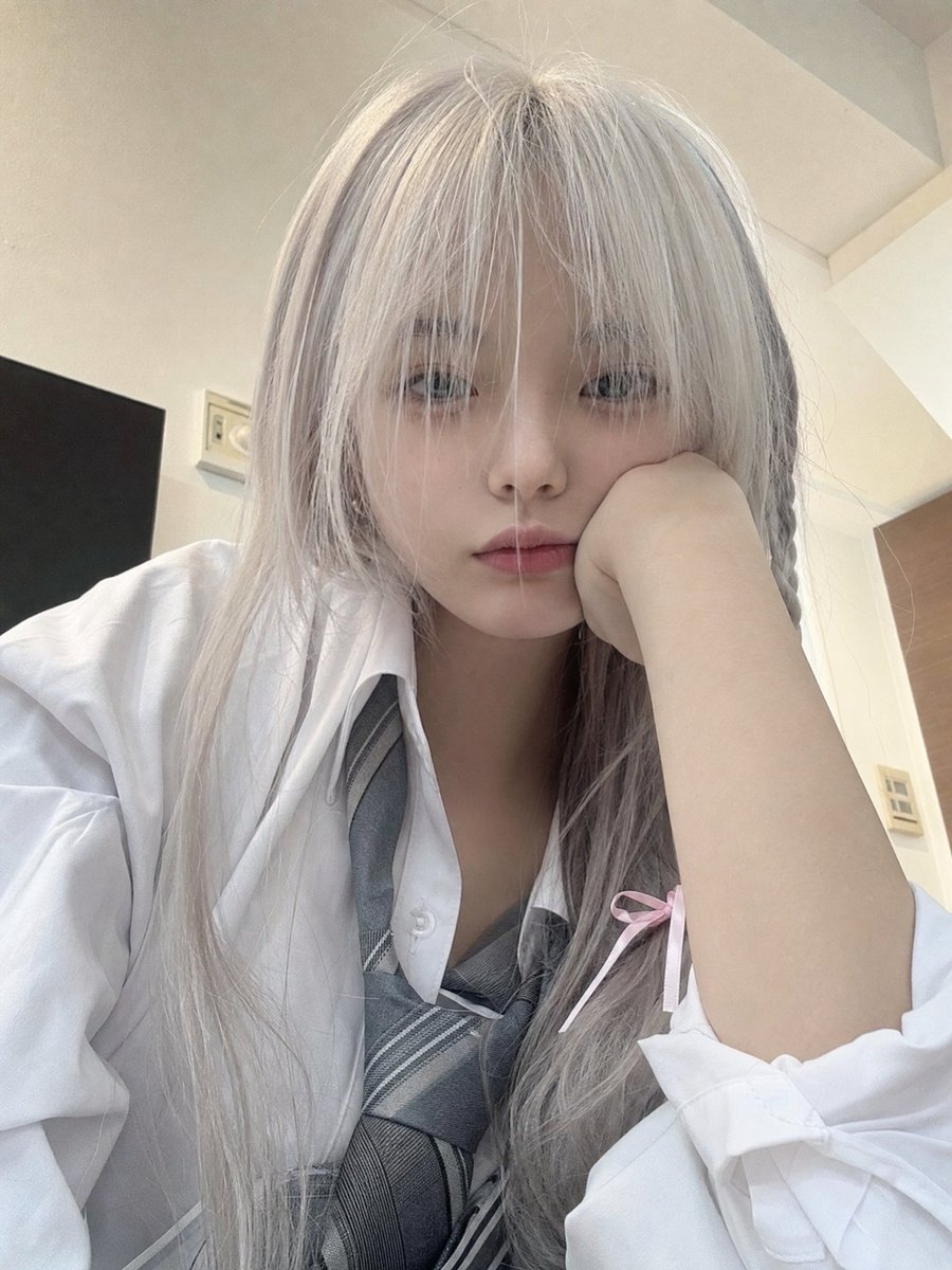 一位年轻的日本女孩,留着长长的银白色头发,柔软凌乱的刘海部分遮住眼睛,侧面编着一条用粉色丝带扎住的小辫子,穿着白色纽扣校服衬衫、灰色领带和校服。她随意地坐在室内,一只手托着脸,用…