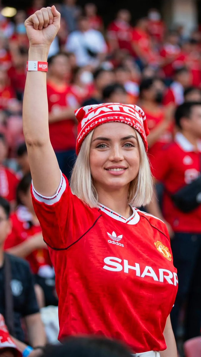 超现实的体育生活方式编辑风格人像：一位惊艳的女性足球迷，留着利落的铂金金色波波头发型，眼神呈冰蓝偏绿色，皮肤纹理自然，妆容柔和，神态自信。她身穿复古款红色曼联…