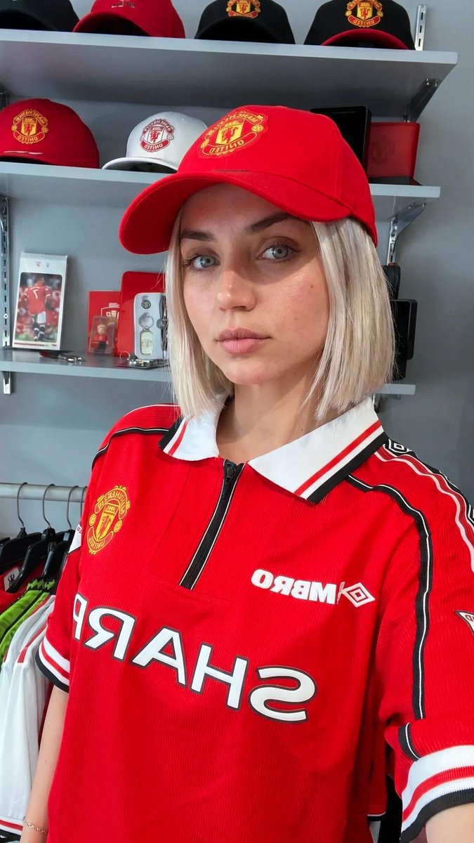 超现实的体育生活方式编辑风格人像:一位惊艳的女性足球迷,留着利落的铂金金色波波头发型,眼神呈冰蓝偏绿色,皮肤纹理自然,妆容柔和,神态自信。她身穿复古款红色曼联球衣(1990年代…