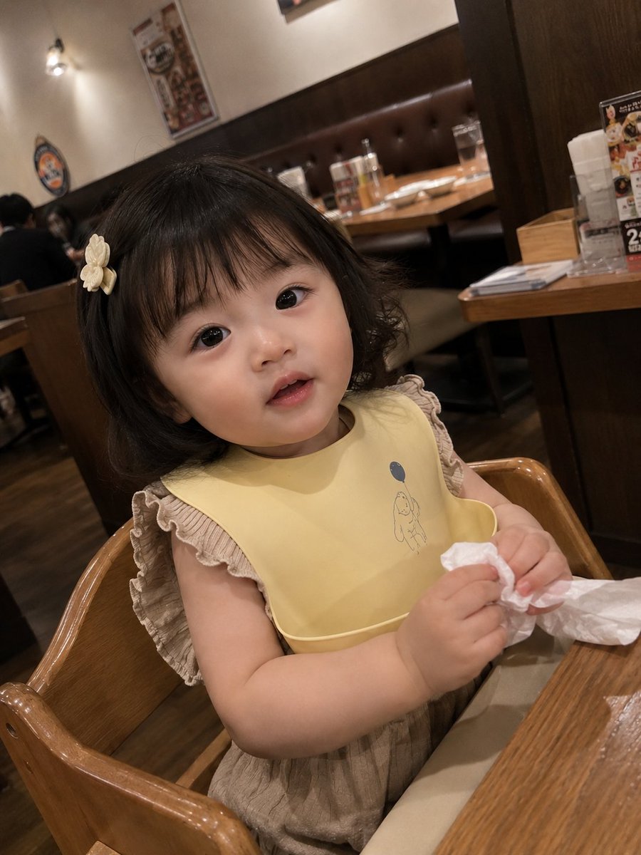 真实感抓拍智能手机照片：一位可爱的女幼儿坐在一家温馨的家庭餐厅的木制高脚椅上。她有着柔软的刘海短发，戴着一个小小的奶白色发夹，眼睛又大又有神，脸颊红润白皙。她身穿浅棕色荷叶边上衣…