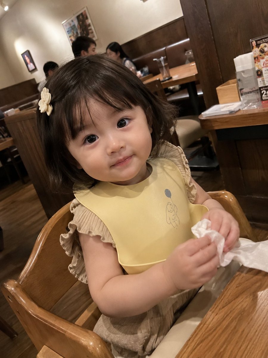 真实感抓拍智能手机照片：一位可爱的女幼儿坐在一家温馨的家庭餐厅的木制高脚椅上。她有着柔软的刘海短发，戴着一个小小的奶白色发夹，眼睛又大又有神，脸颊红润白皙。她身穿浅棕色荷叶边上衣…