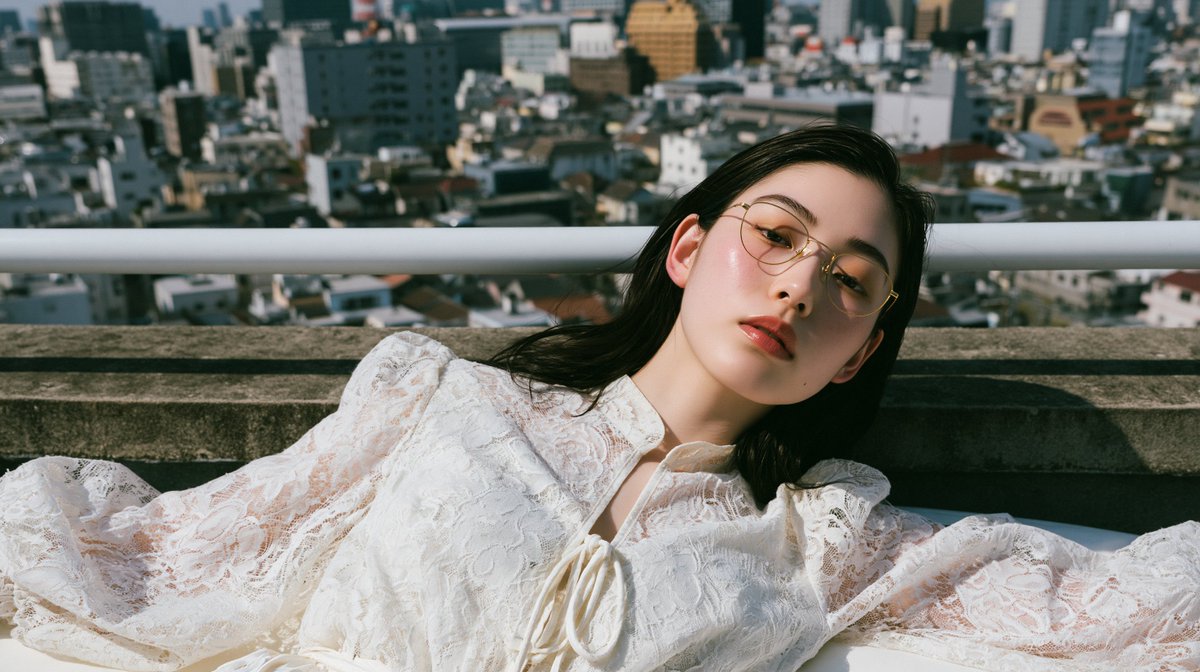 一位惊艳的二十多岁日本或韩国女性，戴着金色金属细框眼镜，身穿白色蕾丝泡泡袖系带领口衬衫，以自然躺卧的姿势斜靠在床沿；头部转向镜头，位于高楼屋顶边缘，背景有屋顶…