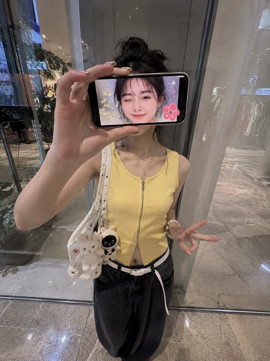 超现实创意iPhone人像照:一位时尚的日本女性站在现代购物中心的反光玻璃墙前,采用略微抬高的正面拍摄角度,呈现0.5x iPhone广角镜头美学。 她横向握着一部智能手机放在脸…