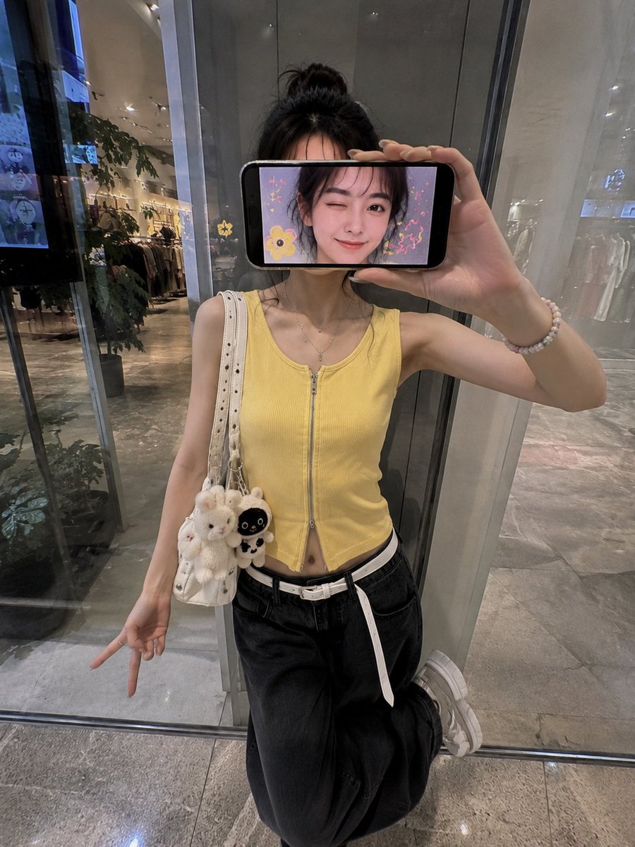 超现实创意iPhone人像照:一位时尚的日本女性站在现代购物中心的反光玻璃墙前,采用略微抬高的正面拍摄角度,呈现0.5x iPhone广角镜头美学。 她横向握着一部智能手机放在脸…