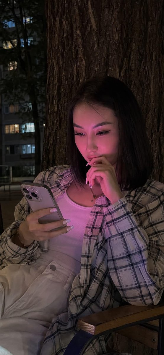 夜晚使用 iPhone 15 Pro 拍摄的写实风格生活照。画面中央是一位年轻女性,留着及腰的黑色长直发。她正饶有兴趣地注视着智能手机,嘴角轻轻上扬,温柔地微笑。 妆容:长簇式睫…