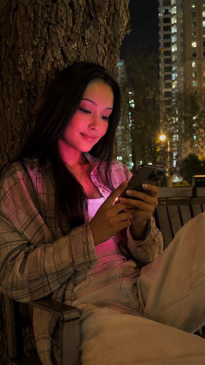 夜晚使用 iPhone 15 Pro 拍摄的写实风格生活照。画面中央是一位年轻女性,留着及腰的黑色长直发。她正饶有兴趣地注视着智能手机,嘴角轻轻上扬,温柔地微笑。 妆容:长簇式睫…