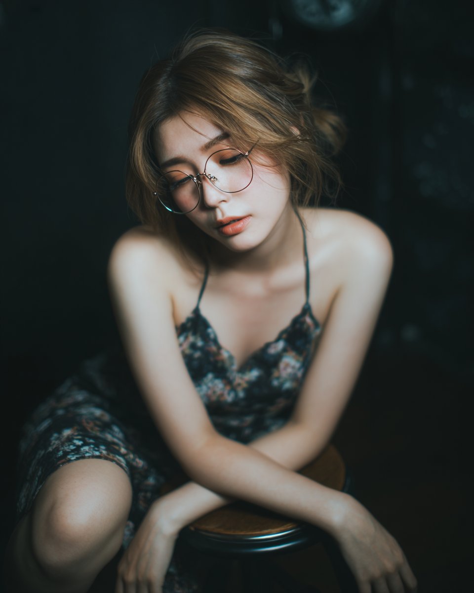 一位二十多岁的迷人日本或韩国女性，戴着金色金属细框眼镜，身穿长裙，以坐姿安排坐在吧台凳上，一膝屈起、另一条腿自然放松；头部低垂朝双手方向，身处近景草地平面之上…