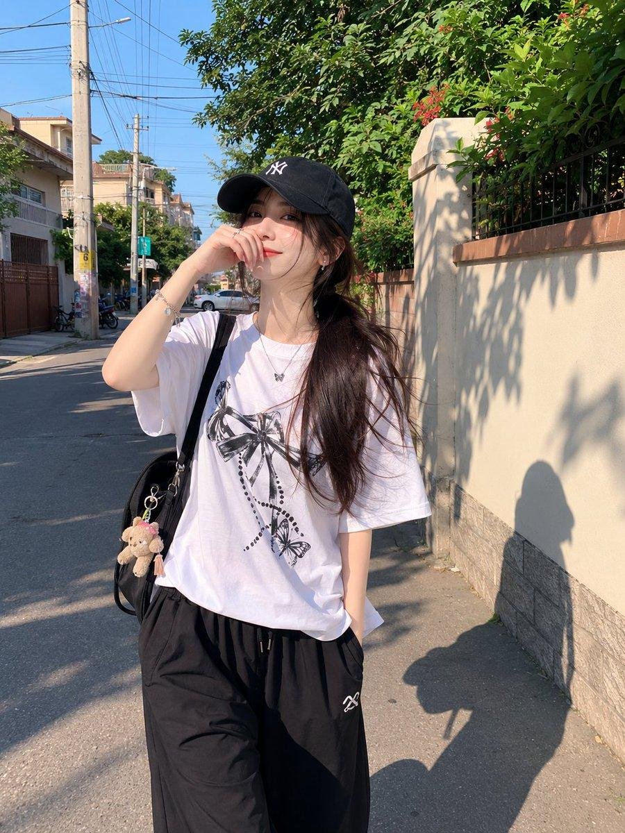 漂亮的年轻日本女孩,肌肤白皙,妆容柔和自然,深色长发在黑色棒球帽下扎成低马尾,身穿宽松的白色印花T恤和黑色宽松全长运动裤。在一个晴朗的午后,她随意地走过一条安静的住宅小巷,一只手…