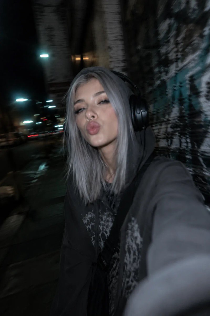 以所附照片为参考，创作一张夜间城市摄影人像，主体为一位年轻女性，整体呈现另类、垃圾摇滚（grunge）美学。她留着齐肩直发、中分，浅色眼睛搭配浓重的黑色猫眼眼线、纤长睫毛，戴着鼻…