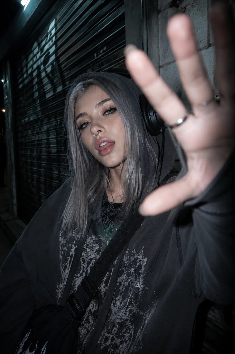 以所附照片为参考，创作一张夜间城市摄影人像，主体为一位年轻女性，整体呈现另类、垃圾摇滚（grunge）美学。她留着齐肩直发、中分，浅色眼睛搭配浓重的黑色猫眼眼线、纤长睫毛，戴着鼻…
