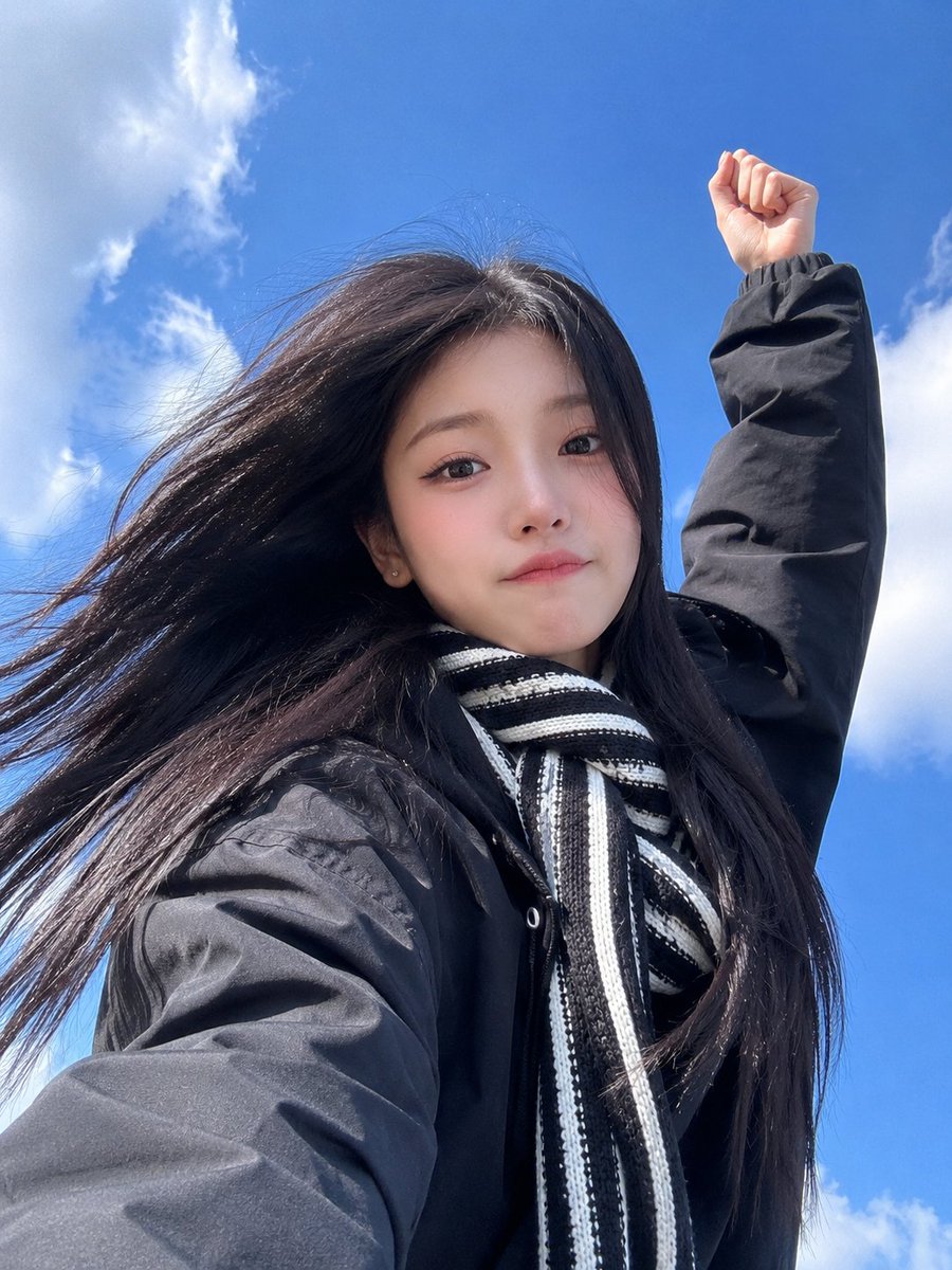 极致真实的智能手机自拍照，一位美丽的东亚年轻女性，从极具戏剧性的低角度仰拍，背景是鲜艳的蓝天和柔和的白色云朵。她拥有又长又直的乌黑长发在风中自然飘动，白皙的瓷器肌肤，柔和的杏仁形…