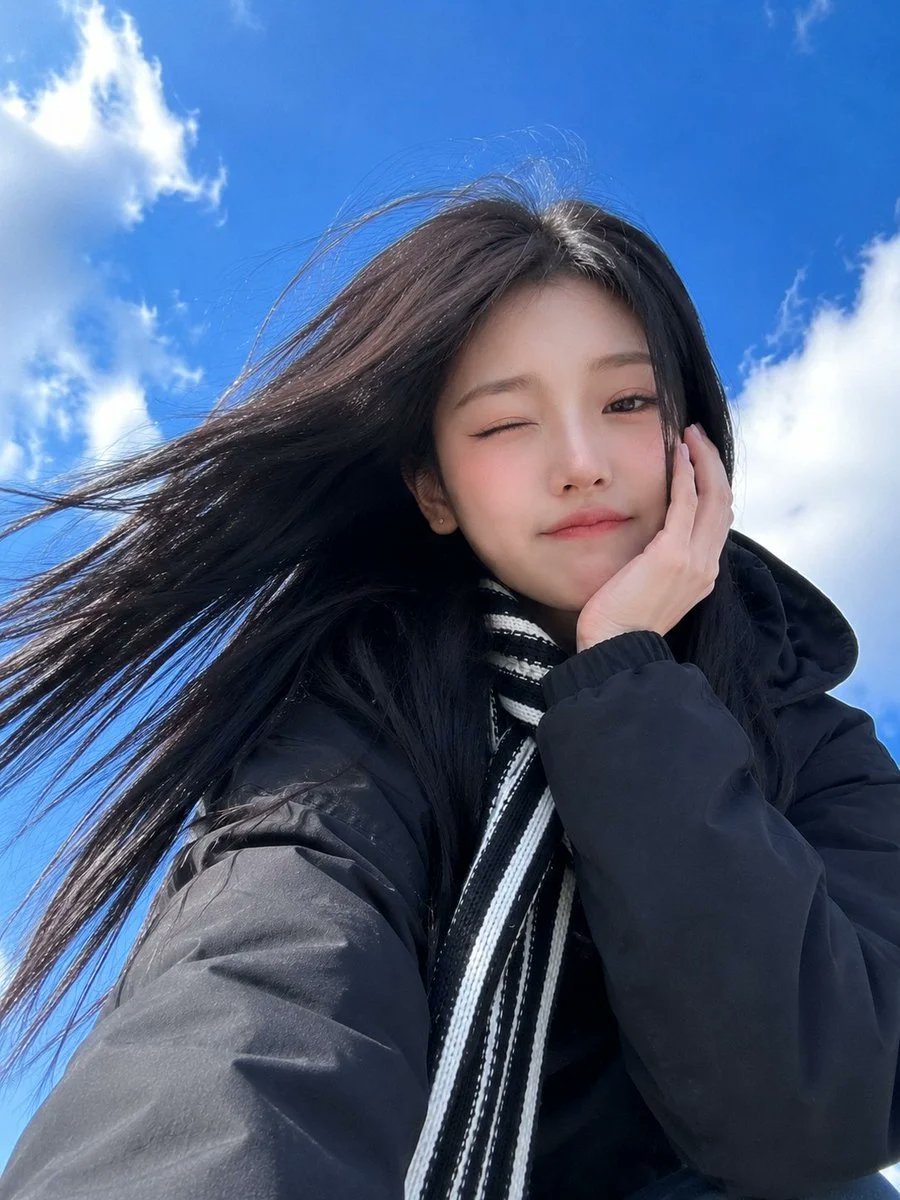 超写实智能手机自拍：一位年轻漂亮的东亚女性，以戏剧性低角度视角拍摄，背景是带有柔软白云的鲜艳蓝天。她留着顺直乌黑长发，在风中自然飘动，拥有白皙瓷肌、柔和的杏仁形棕眼、细腻的翼状眼…