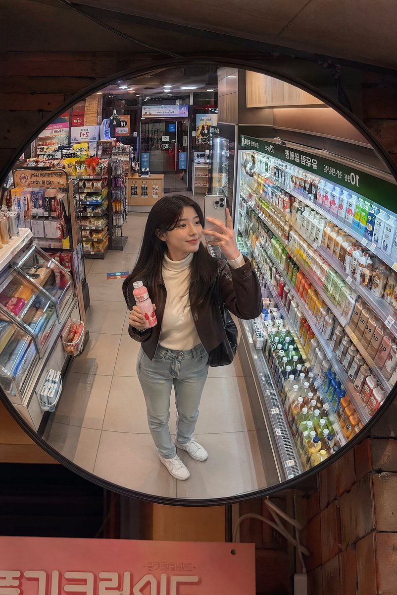 深夜韩国便利店镜面抓拍 创建一张逼真的生活方式照片，透过安装在韩国一家温馨便利店天花板上的一面大型凸面镜拍摄，场景为夜晚。画面中一位时尚的年轻韩国女性从略微抬高的镜面视角被捕捉到…