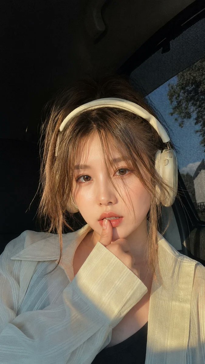黄金时段坐在豪华轿车内的美丽年轻女子的超写实特写肖像,佩戴优雅的米白色头戴式耳机。柔和的自然阳光从侧面车窗洒入,在她的脸庞和宽松的白色衬衫上投射出温暖的电影般的高光。她拥有白皙的…