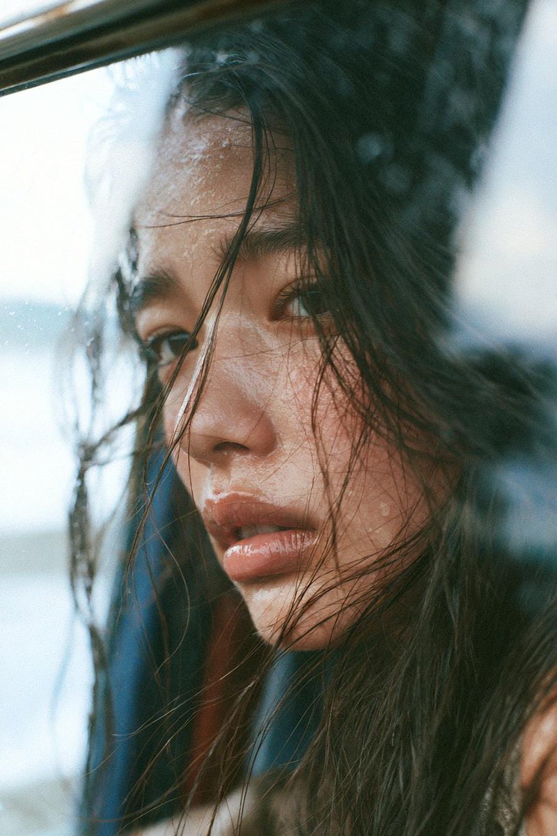 一位忧郁的年轻东亚女性,留着略显凌乱的长黑发,在黄金时刻静静坐在一扇大窗旁,柔和的阳光以温暖的电影色调照亮她的面庞。她若有所思地望向远方,妆容自然,皮肤水润,眼神富有表现力,几缕…