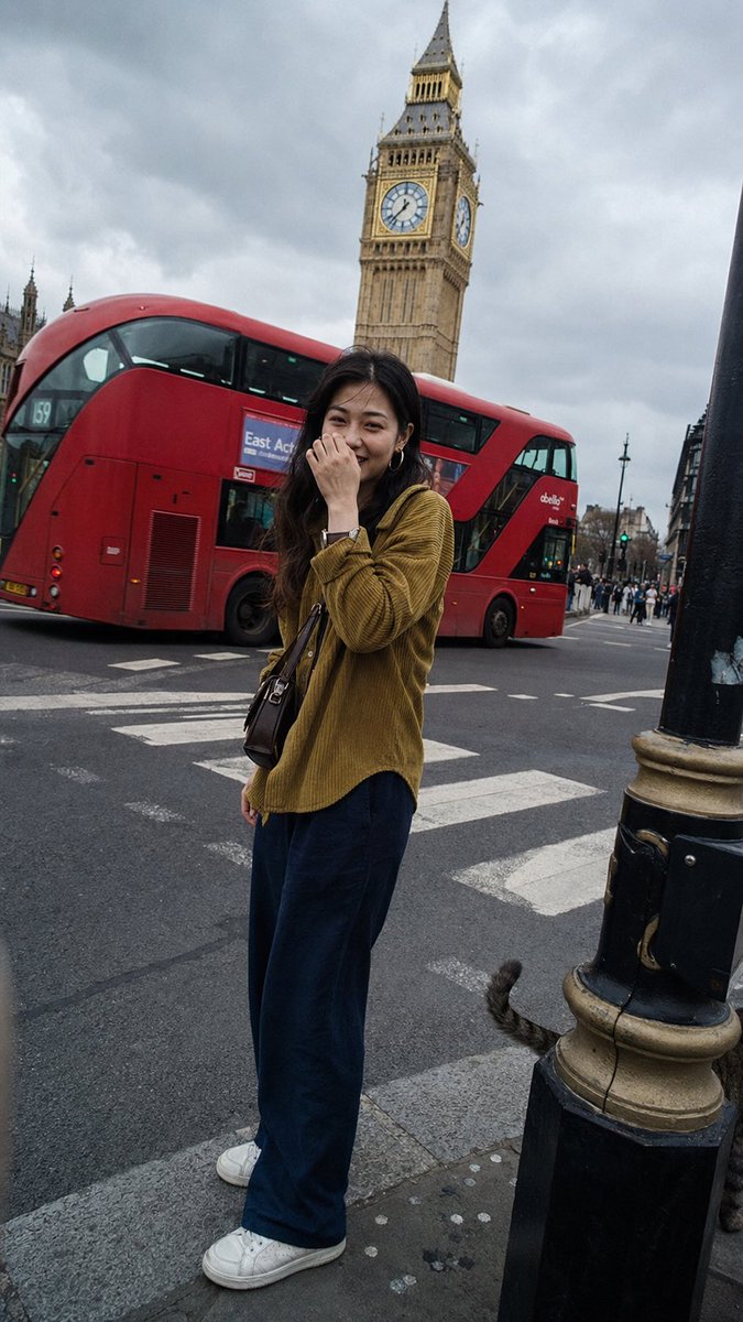 一张在伦敦街头随手拍摄的普通抓拍快照，看起来像是有人走在她前面，转身拍了一张照片。一位二十出头的年轻东亚女性站在人行横道前，她的容貌百分之百写实——直视镜头…