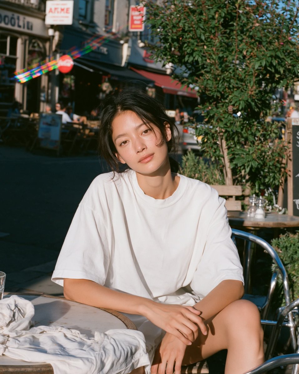 温暖阳光下的胶片人像，年轻女性放松地坐在小咖啡馆露台桌旁沐浴在明亮的金色午后阳光下，目光平静柔和、双眼睁开，身穿超大号白色T恤，身后是清晰可辨的户外街景，场景明亮通透，一束棱镜般…