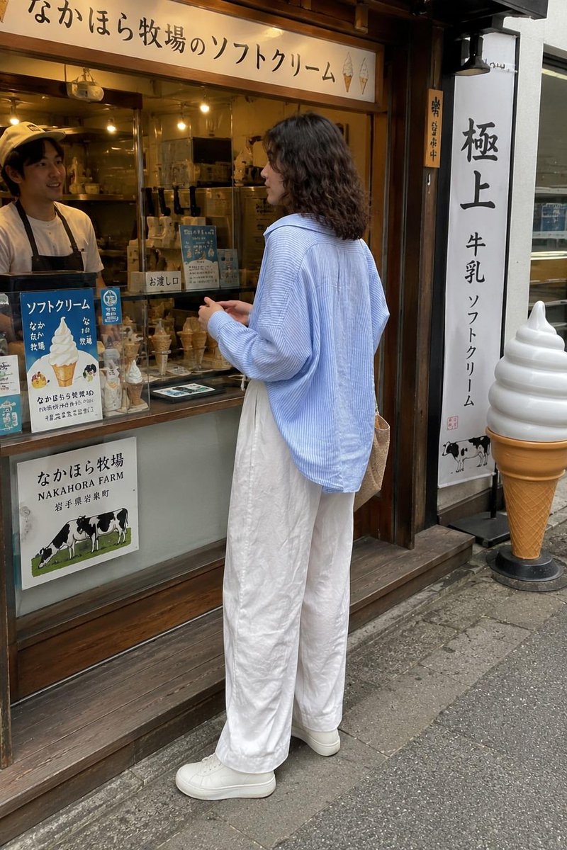 一张抓拍的旅行照片：一位年轻女子站在一家传统日式软冰淇淋店外，回眸对镜头微笑。她留着齐肩深色波浪卷发，身穿宽松的浅蓝色条纹纽扣衬衫、宽松的白色阔腿亚麻长裤和白色极简风格运动鞋，手…