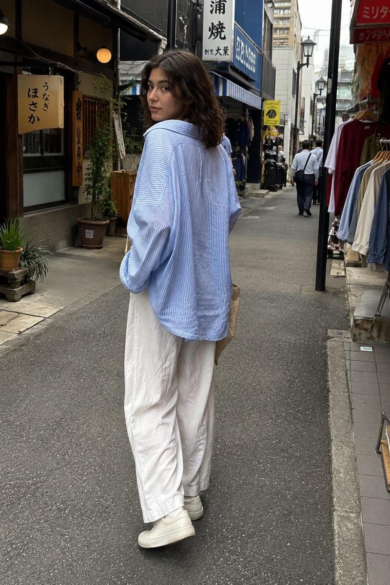 一张抓拍的旅行照片：一位年轻女子站在一家传统日式软冰淇淋店外，回眸对镜头微笑。她留着齐肩深色波浪卷发，身穿宽松的浅蓝色条纹纽扣衬衫、宽松的白色阔腿亚麻长裤和白色极简风格运动鞋，手…