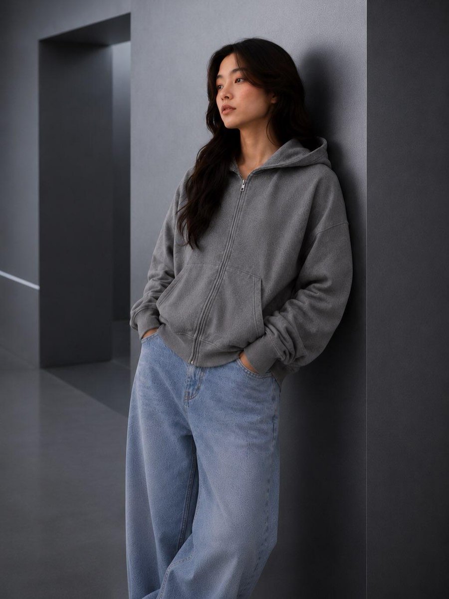 极简时尚编辑人像：一位年轻女性随意倚靠在一面光滑的炭灰色建筑墙面上，身穿水洗灰色宽松拉链连帽衫、浅色水洗宽松牛仔裤和白色运动鞋，双手插兜，姿态放松，神情平静自…