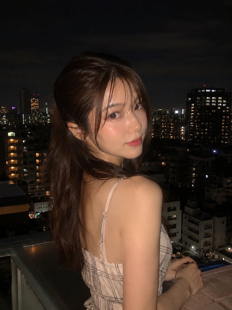 超写实夜景人像:一位美丽的年轻女子站在阳台上,俯瞰城市天际线,身穿优雅的无袖米色格纹连衣裙,淡雅腮红妆容,自然水润唇妆,慵懒的半扎发造型,几缕碎发自然垂落于脸颊两侧。她回眸望向镜…