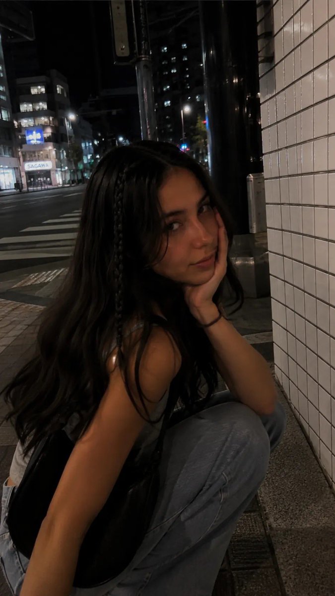 一段电影感十足的夜间街拍人像，写实抓拍风格，在日本城市安静的人行道上用 iPhone 拍摄。一位年轻女性沿着人行道行走，长而微卷的深棕色头发，一侧脸旁垂着一根细辫子，妆容自然柔和…