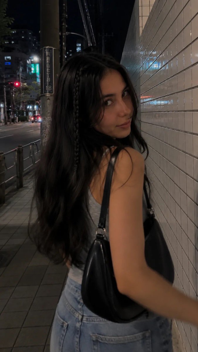 一段电影感十足的夜间街拍人像，写实抓拍风格，在日本城市安静的人行道上用 iPhone 拍摄。一位年轻女性沿着人行道行走，长而微卷的深棕色头发，一侧脸旁垂着一根细辫子，妆容自然柔和…
