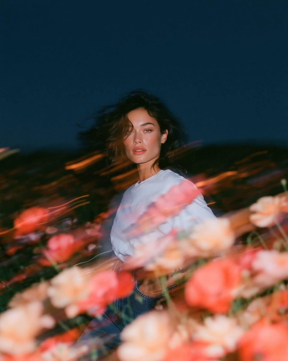 写实风格的实验性时尚人像:一位天生丽质的年轻女子正面站立于蓝色时刻狂风拂过的野花田中,镜头从大腿向上以平视取景。她身穿一件宽松的纯白棉质T恤,搭配水洗中蓝色直筒牛仔裤,日常随性的…
