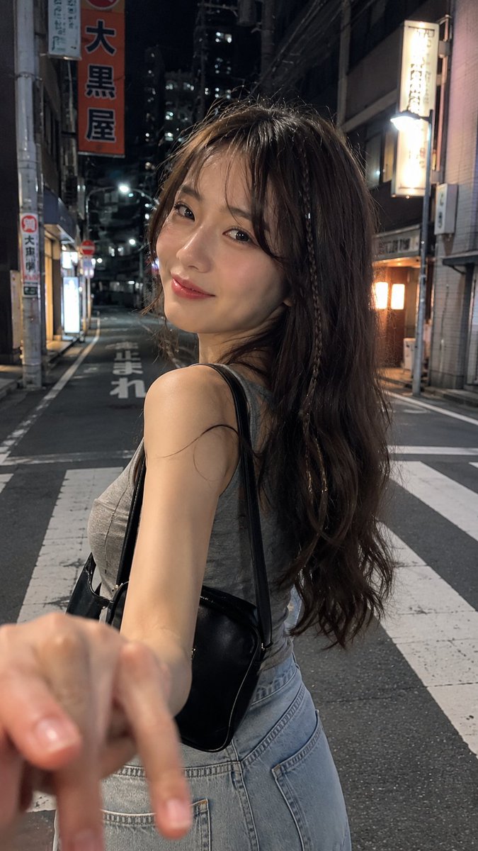 一张电影感十足的夜间街头抓拍人像，拍摄于日本街头，使用 iPhone 拍摄。一名年轻女子，长长的深棕色波浪卷发中编有一缕细辫，淡妆，嘴角带着若有若无的微笑，回…