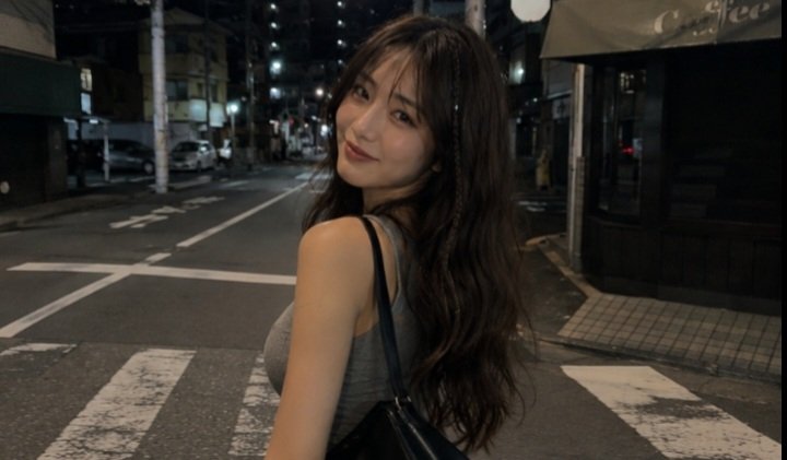 一张电影感十足的夜间街头抓拍人像,拍摄于日本街头,使用 iPhone 拍摄。一名年轻女子,长长的深棕色波浪卷发中编有一缕细辫,淡妆,嘴角带着若有若无的微笑,回头望向镜头。她身穿灰…