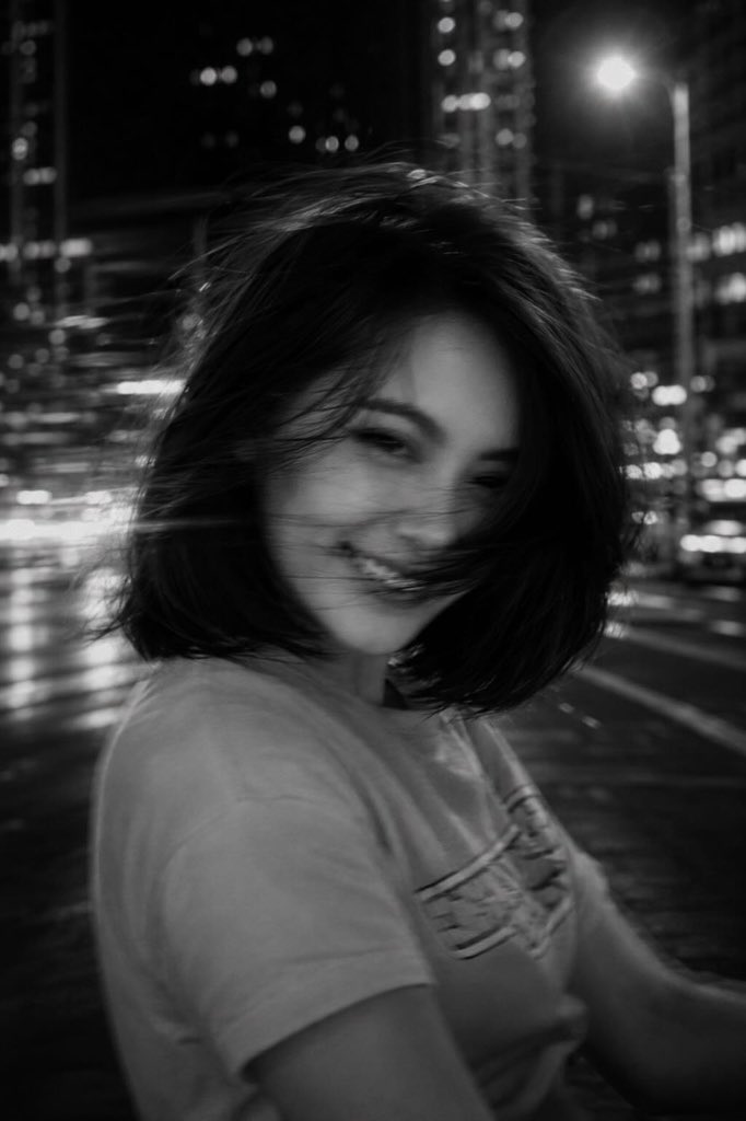 一张黑白电影感的城市夜景照片,画面中一位年轻女子位于前景,略微偏离中心,正在转头过程中,长发在画面中甩动成柔和、幽灵般的发丝。长曝光拍摄,带有意运动模糊。她面部特征部分模糊但仍可…