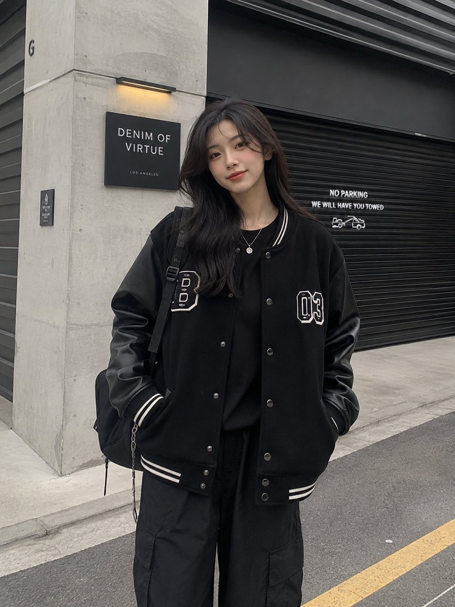 一张潮流街头服饰时尚人像照：一位时尚年轻女性自信地站在一座现代工业建筑前，建筑拥有混凝土墙面和一扇黑色金属卷帘门。她身穿一件超大版型的黑色棒球外套，袖部为皮革…