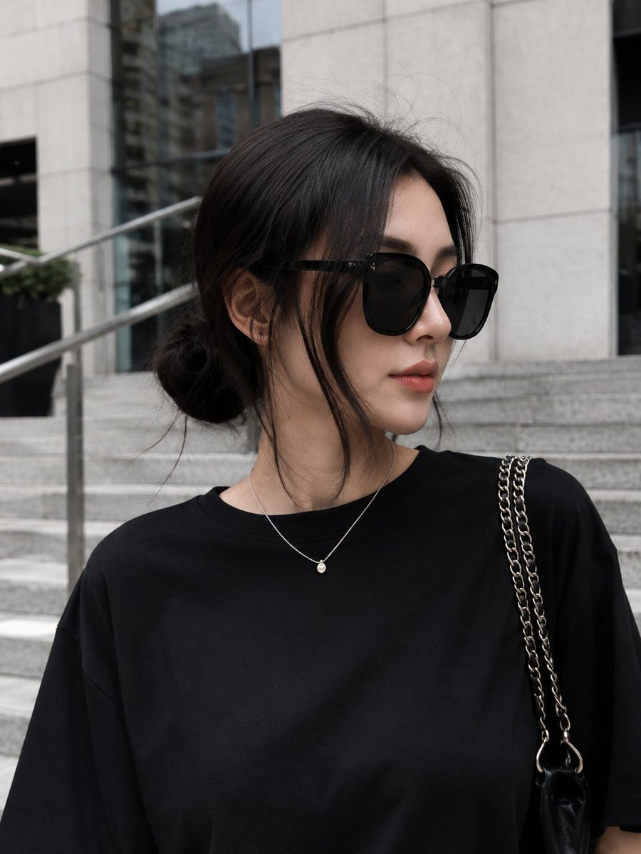 极简奢华街头风格时尚人像:一位时尚年轻女性自信地走下一座当代建筑外的宽阔现代花岗岩石阶。她身穿超大号黑色T恤,搭配宽松的黑色阔腿裤,脚踩清爽的白色运动鞋,佩戴超大黑色太阳镜,肩背…