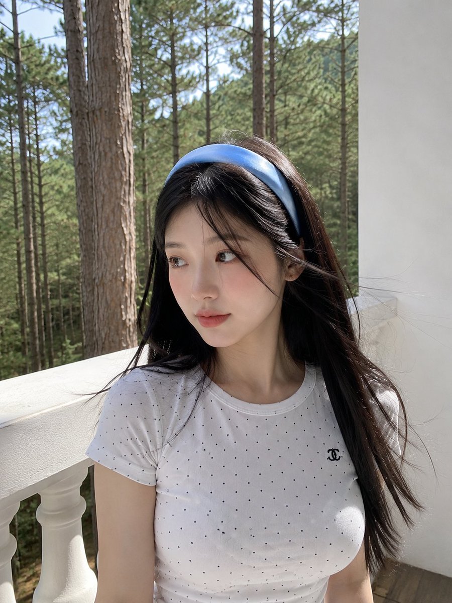 超写实的智能手机照片：一位美丽的年轻日本女性站在被宁静松林环绕的白色阳台上。她拥有白皙的瓷器肌肤、柔和的玫瑰色脸颊、深棕色的大眼睛、精致细腻的五官，以及在微风中轻轻飘动的黑色长直…
