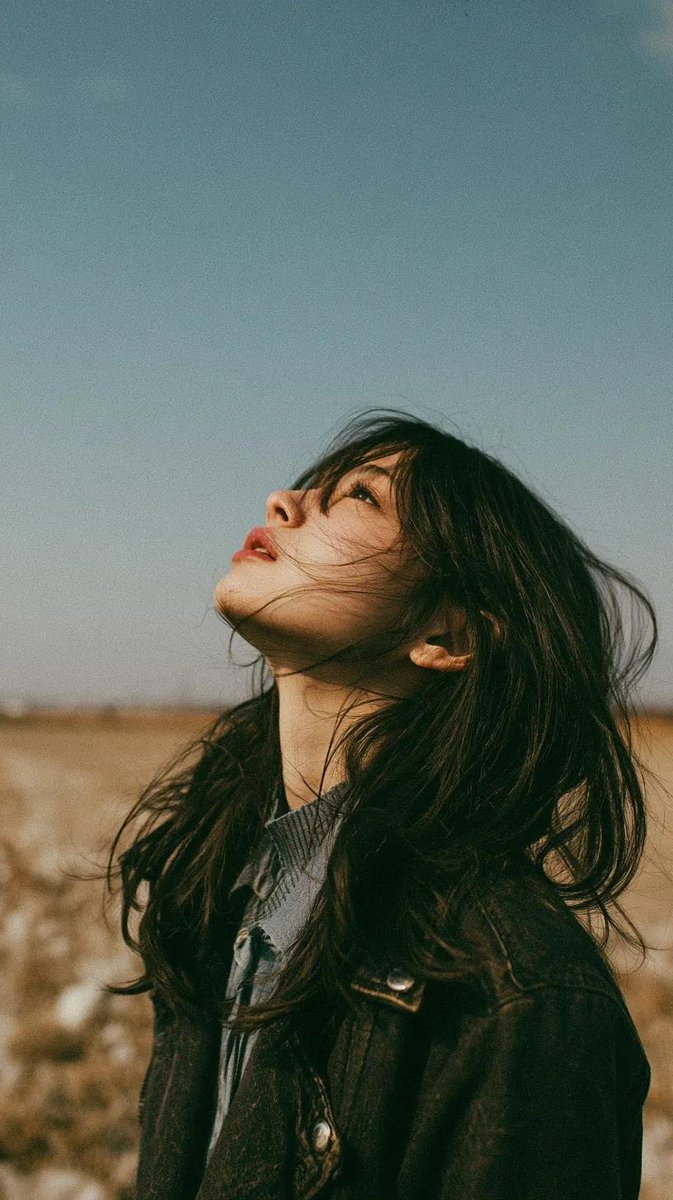 电影极简风格肖像：一位美丽的年轻东亚女性独自伫立在开阔的野花草甸上，头顶是无限的蓝天，她双眼微闭或半睁，仰望天空，仿佛在拥抱微风。乌黑长发自然蓬松，随风自由飘…