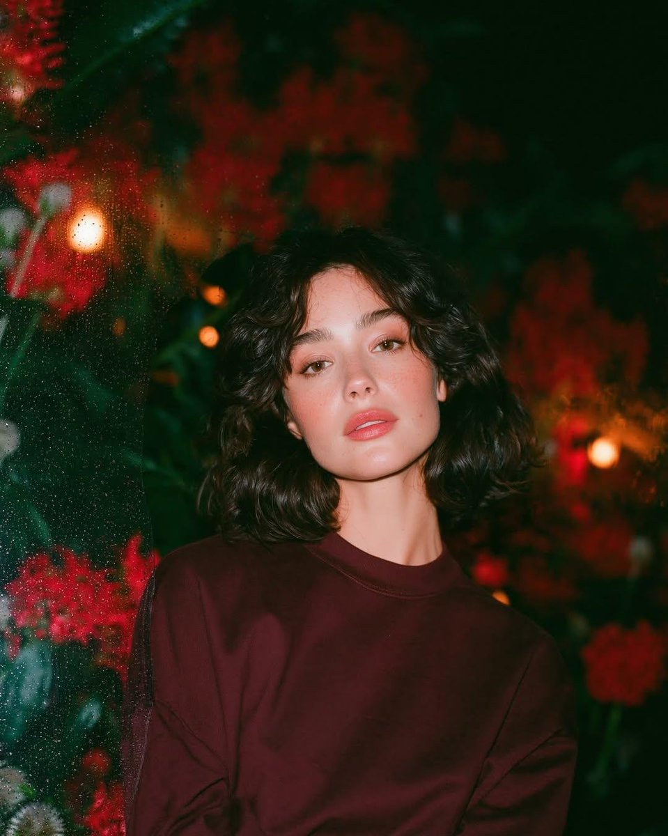 梦幻的电影感夜景人像：一位美丽的年轻女性，留着短款波浪深色头发，皮肤散发着自然光泽，身穿一件宽松的酒红色圆领卫衣，夜晚站在一座郁郁葱葱的花园中，园内开满了鲜艳…