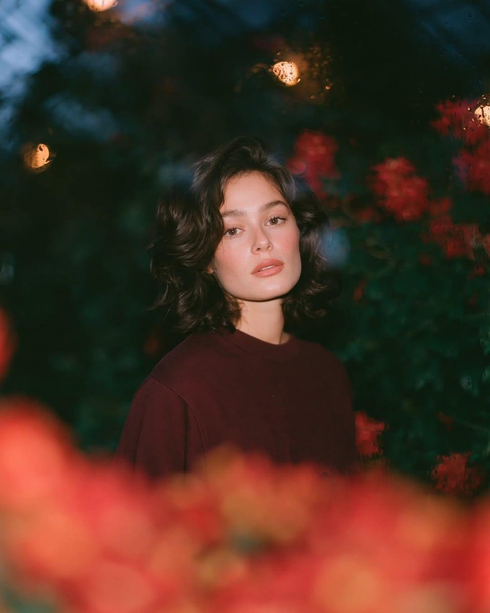 梦幻的电影感夜景人像：一位美丽的年轻女性，留着短款波浪深色头发，皮肤散发着自然光泽，身穿一件宽松的酒红色圆领卫衣，夜晚站在一座郁郁葱葱的花园中，园内开满了鲜艳的红色花朵和深绿色的…