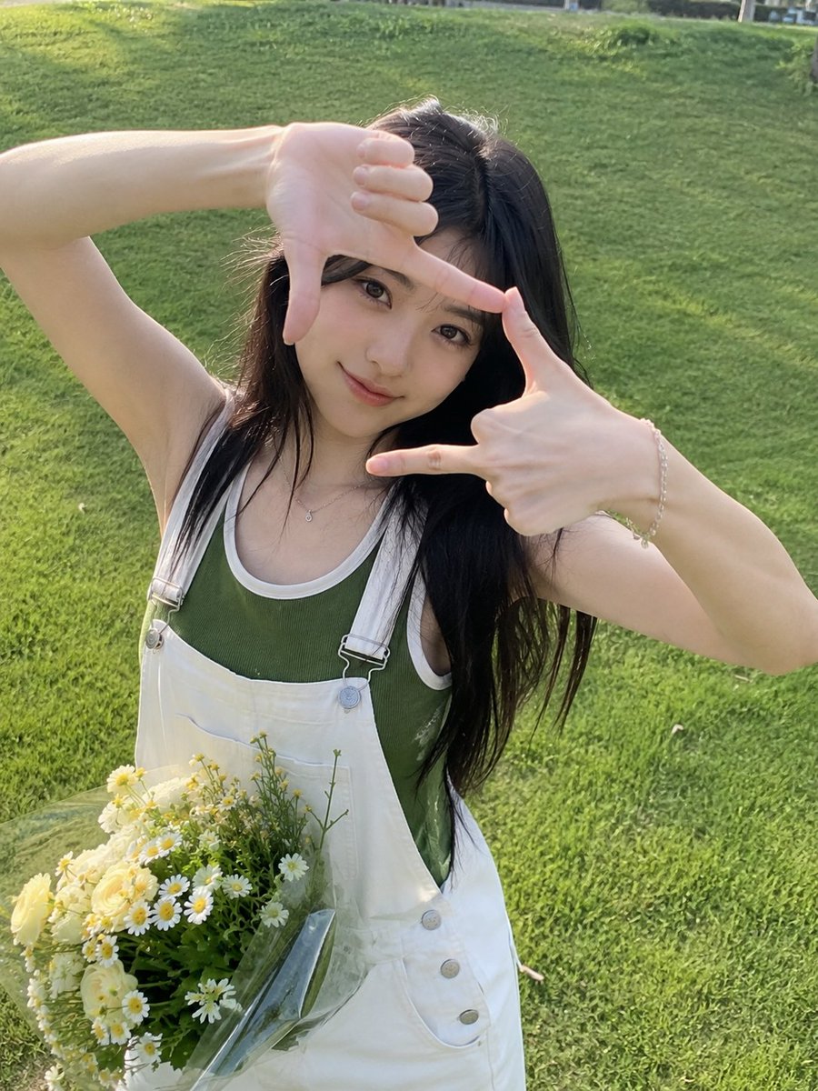 一张迷人的户外人像照,一位年轻女子站在郁郁葱葱的绿色草坪上,直视镜头,双手俏皮地在脸部周围比划出一个长方形相框。她有着一头长而深色、柔和波浪卷的头发,带着温柔…