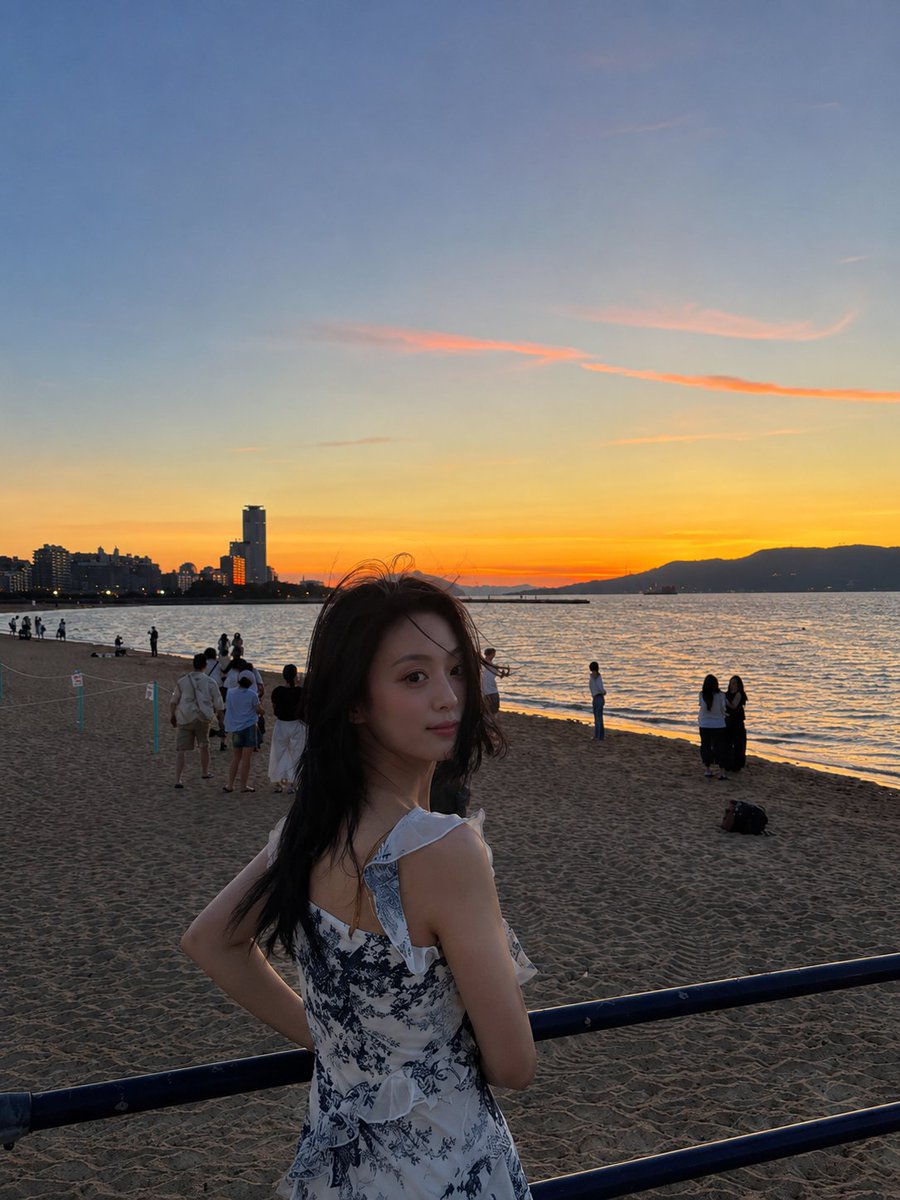 超现实电影感旅行照片：一位年轻的东亚女性站在沙滩上，沐浴在令人屏息的日落中，采用竖向 3:4 构图。她身穿一件精致的白色连衣裙，点缀着细腻的蓝色花卉印花，肩带为荷叶边设计，整体剪…