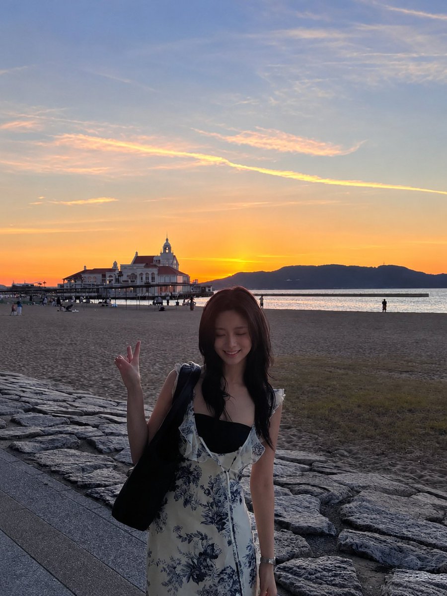超现实电影感旅行照片：一位年轻的东亚女性站在沙滩上，沐浴在令人屏息的日落中，采用竖向 3:4 构图。她身穿一件精致的白色连衣裙，点缀着细腻的蓝色花卉印花，肩带为荷叶边设计，整体剪…