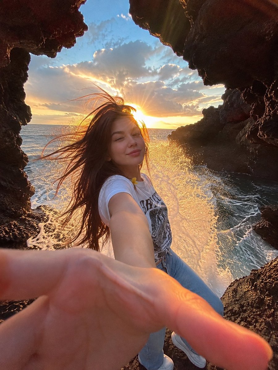一幅电影感十足的海边人像照：一位年轻女子站在天然海洞内的黑色火山岩上，洞口粗犷的岩石形成天然画框。她一头深色长发在海风中戏剧性地飘扬，对着镜头俏皮地眨着一只眼，嘴角带着温柔微笑…
