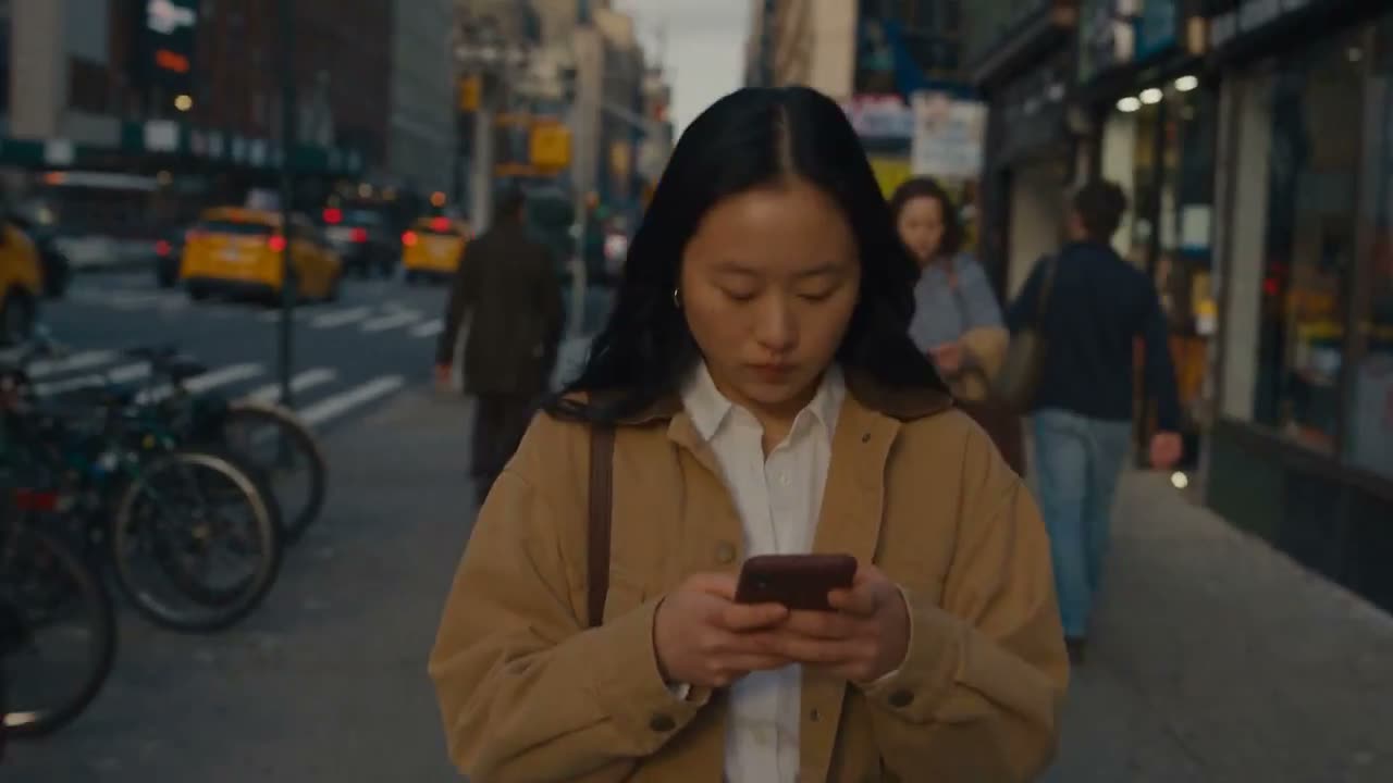 场景 1 — 忙碌的早晨 | 0:00–0:05 "手持摄影的真实感街头画面：一位二十多岁的年轻亚洲女性在清晨走过繁忙的城市人行道。她身穿米色夹克、白色衬衫、…