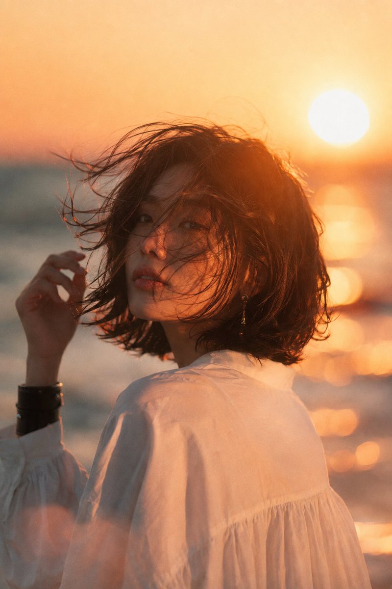 海滩日落中的美丽年轻亚洲女性，中景电影肖像。深色短发波波头在海风中飘扬，头部微向左倾，手正在整理头发。甜美浪漫的瞬间。精致优雅的耳饰，简约宽松的白色衬衫，左腕佩戴黑色宽手镯。柔和…