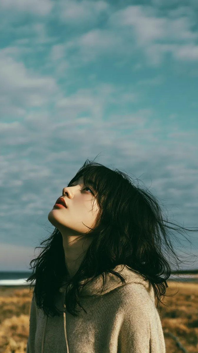 电影极简风格人像：一位美丽的年轻东亚女性独自站立在一片开阔的野花草地上，头顶是无尽的蓝天，她微微仰头，双眼或闭或半睁，仿佛在拥抱风。黑色长发自然蓬松、随风自由拂过面庞，肌肤柔润如…